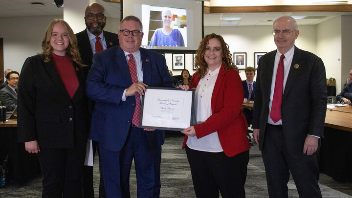 Sheila Stewart appears via Zoom on a screen in the background as university leaders hold her Kudos award during the University of Nebraska Board of Regents meeting on Feb. 9.