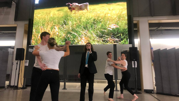 Dancers move around a person singing in the center with an image of a field projected behind them.